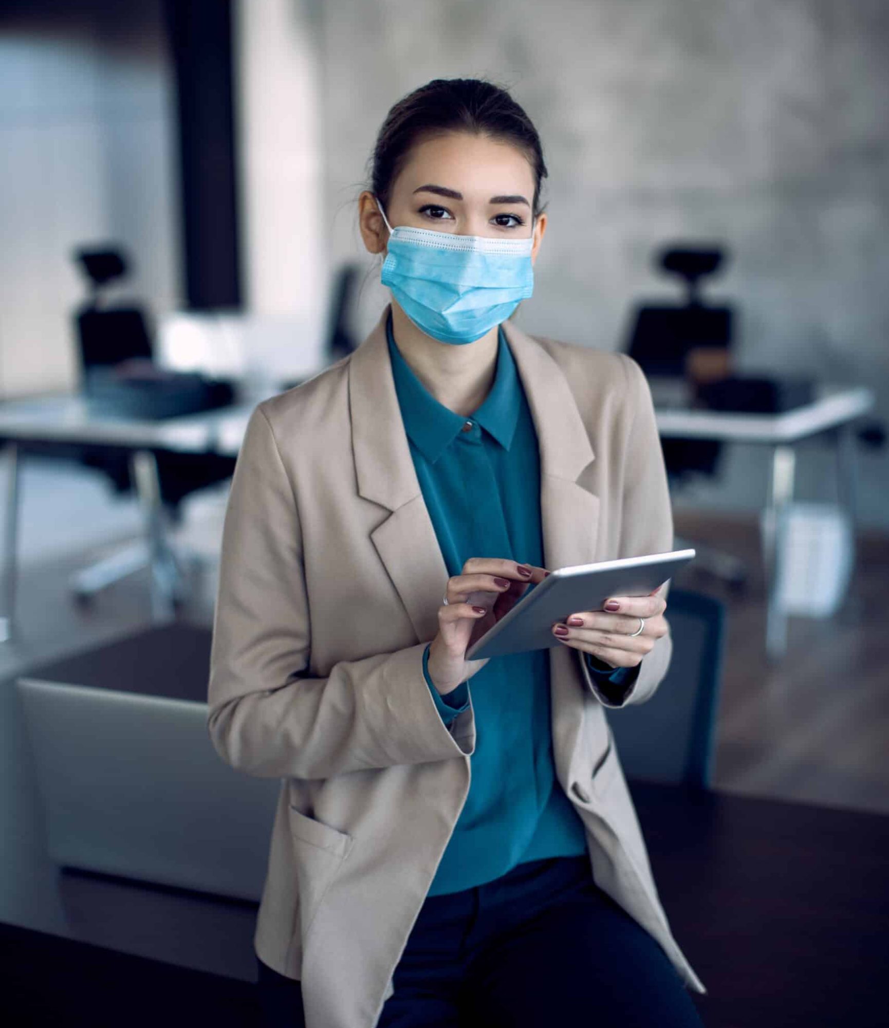 Young businesswoman using touchpad while wearing face mask in the office and looking at camera.